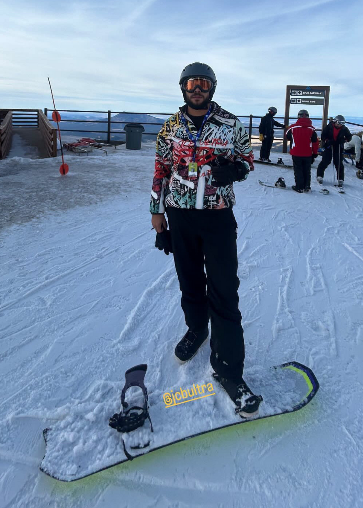 Person with a snowboard on a snowy slope with other snowboarders in the background, wearing the city chaos pullover by jcb ultra